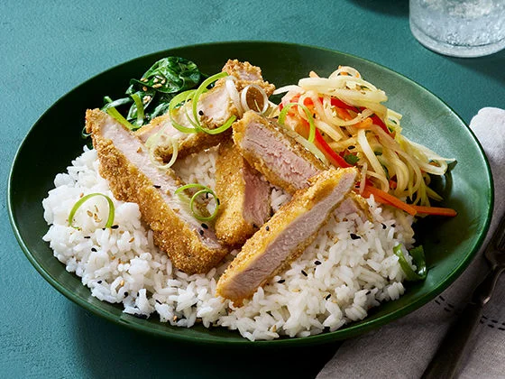 Slices of pinipig-coated pork chop on a bowl of white rice with papaya slaw, baby spinach, scallion curls and sesame seeds.