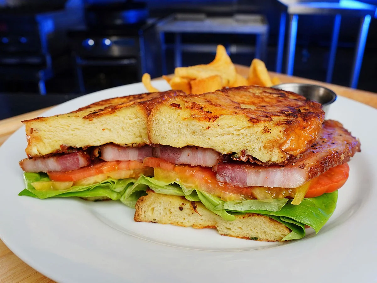 Candied bacon BLT on Parmesan French toast with tomatoes and lettuce, served with fries in a kitchen setting