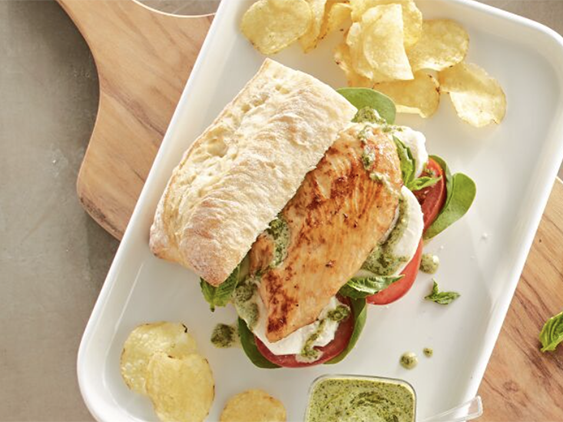 Grilled chicken sandwich with mozzarella, tomatoes, spinach, basil, and pesto aioli on French bread, served on a white plate