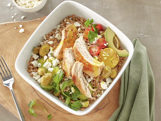 Chicken Breast Filet is served on a bed of farro, topped with feta cheese, avocado, cucumber, tomato and olives