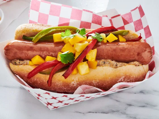 Island Dog with Cheddarwurst, peppers, mango, pineapple, jerk aioli, and cilantro in a tray on marble