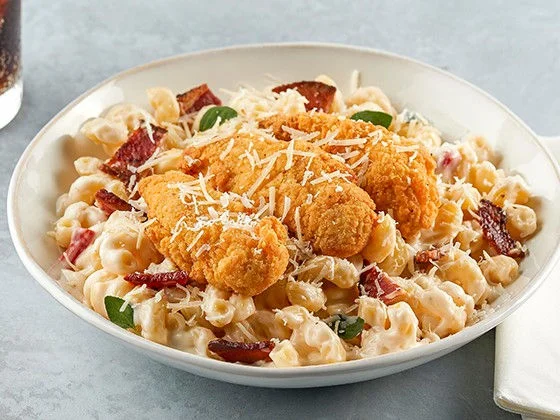 Mac and cheese topped with chicken tenders, bacon, Parmesan, and sliced scallions in a white bowl