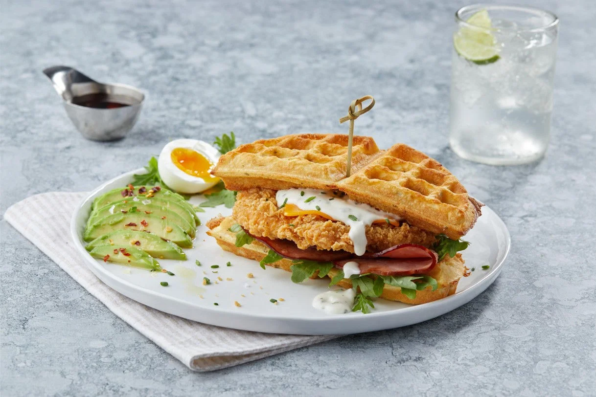 chicken waffle sandwich on a plate with a side of sliced avocado and a soft boiled egg.