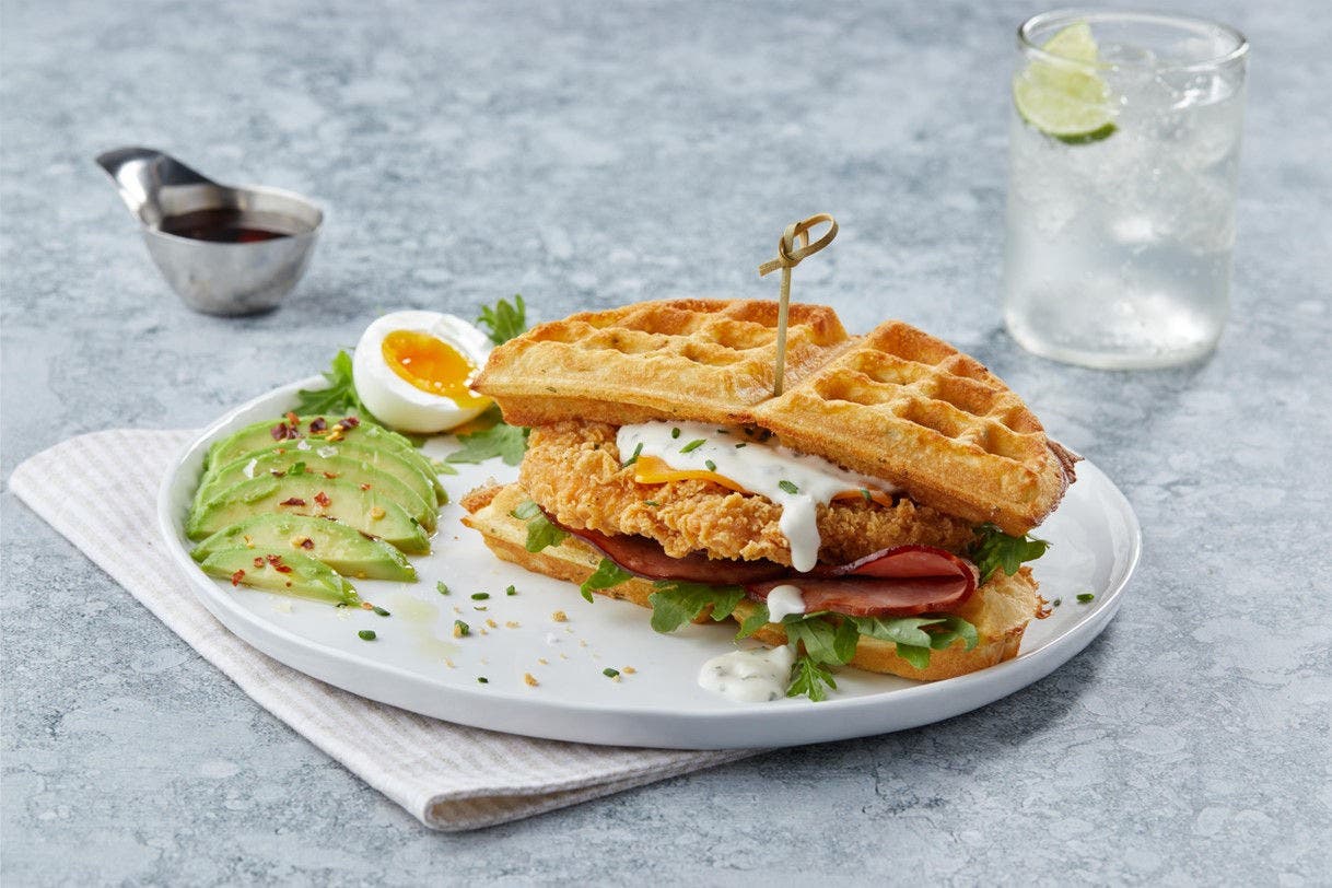 chicken waffle sandwich on a plate with a side of sliced avocado and a soft boiled egg.