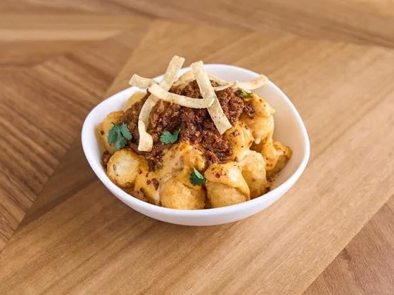 A bowl full of seasoned tots topped with beef taco meat, nacho cheese, creamy caliente Aji Amarillo sauce, cilantro and crispy tortilla strips.
