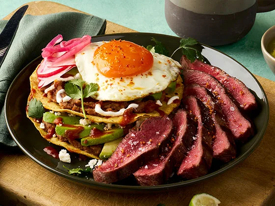 Grilled steak with a tostada topped with beans, avocado, egg, queso fresco, and pickled onions on a dark plate